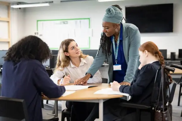 Waist-up view of 15 and 16 year old classmates in uniforms sitting at round table taking notes and discussing ideas.